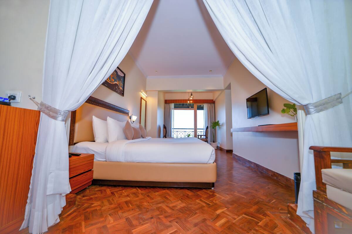 A bedroom with a large bed, wooden flooring, white curtains, a TV on the wall, and a balcony with plants.