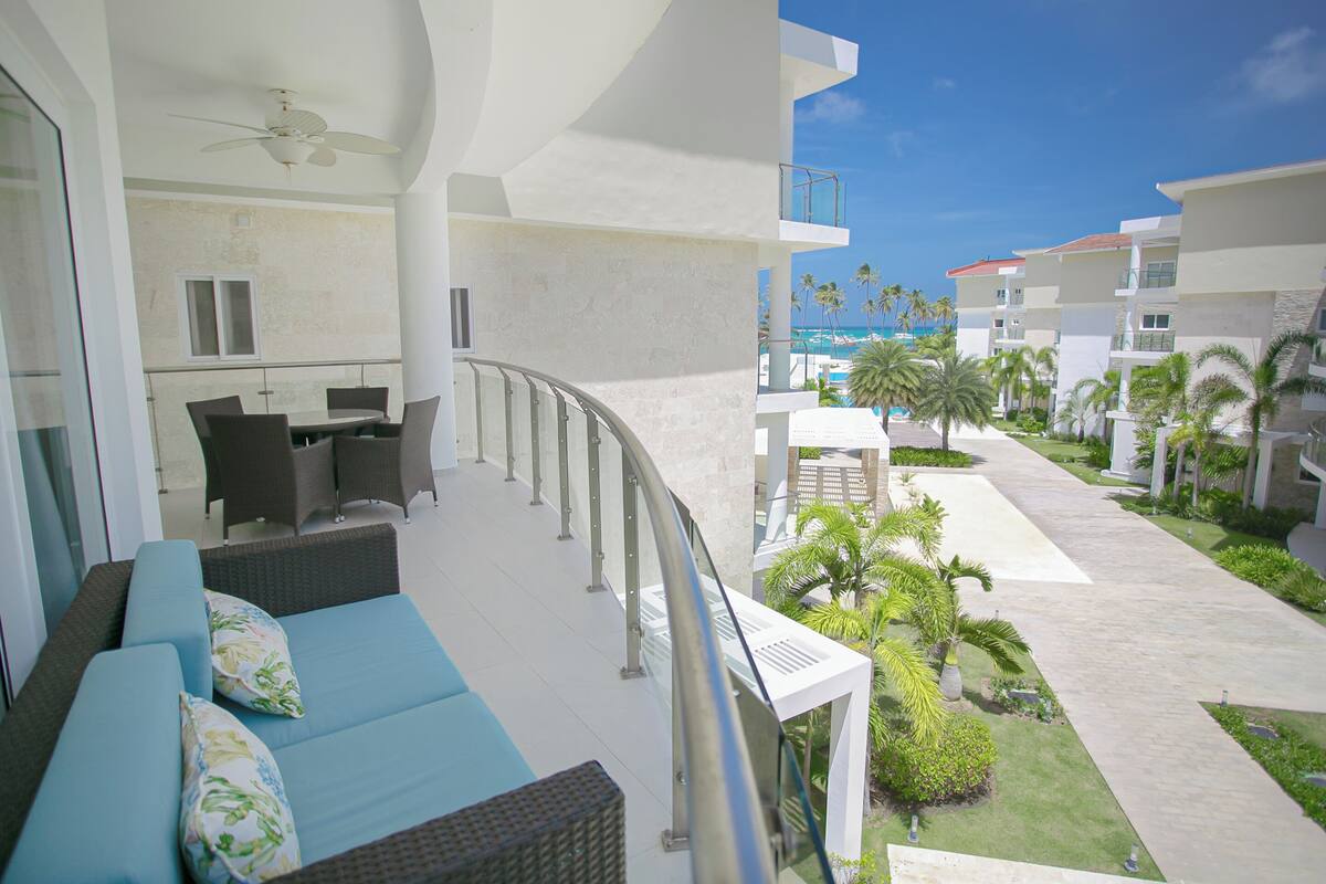 Un balcon avec un canapé et des fauteuils, un espace piscine agrémenté de palmiers et une vue dégagée sur l’océan.