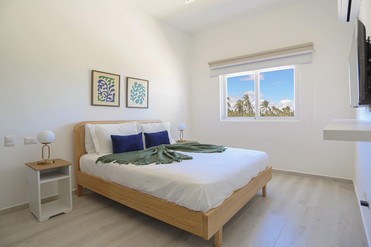 Une chambre avec un lit en bois, une literie blanche, deux oreillers, un plaid vert, une table de chevet avec une lampe et une fenêtre donnant sur des palmiers.