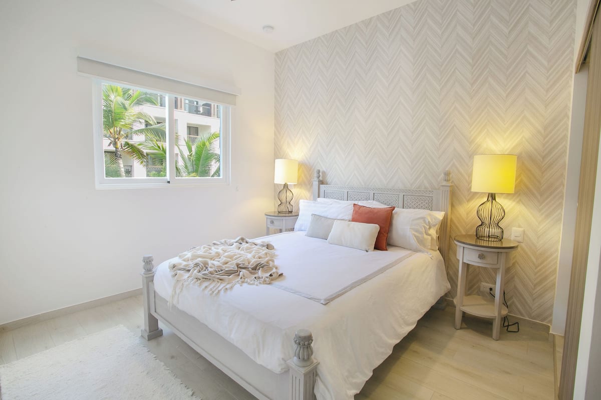 Une chambre à coucher avec un lit, deux tables de chevet avec des lampes, une fenêtre donnant sur des palmiers et un mur orné d’un motif en chevrons.