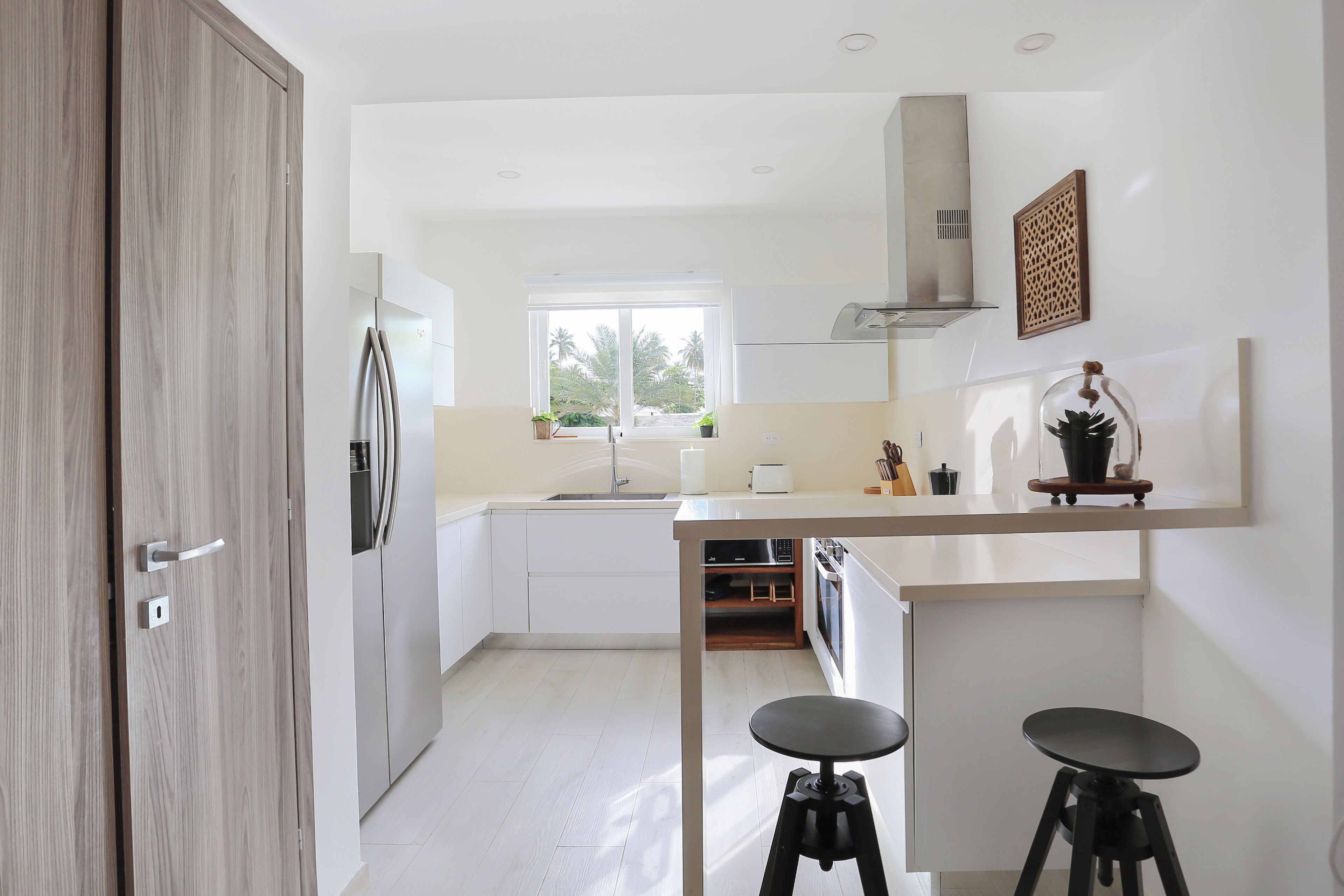 Una cocina moderna con armarios blancos, un refrigerador de acero inoxidable, una ventana con vista a follaje y dos taburetes en una encimera.