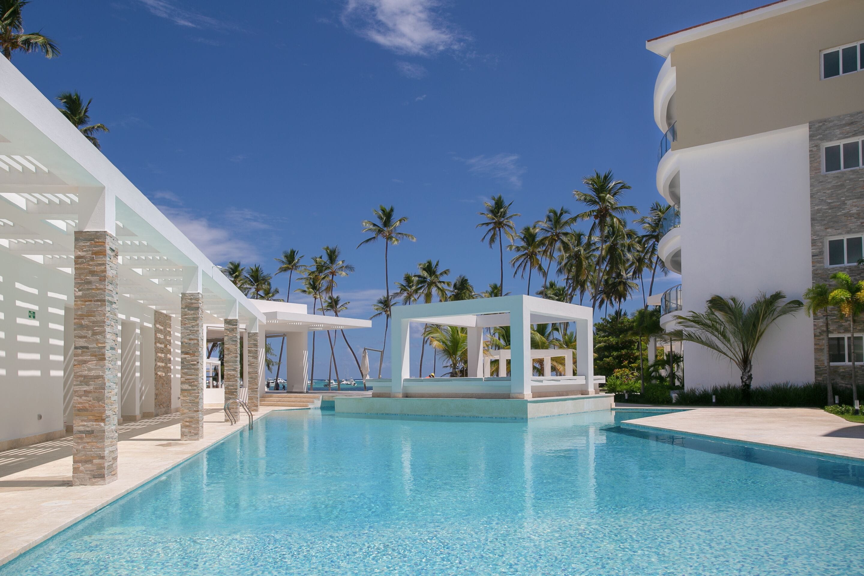 A pool area with clear blue water, palm trees, and a modern building with a white facade and stone pillars.
