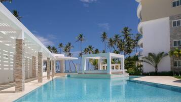 A pool area with clear blue water, palm trees, and a modern building with a white facade and stone pillars.
