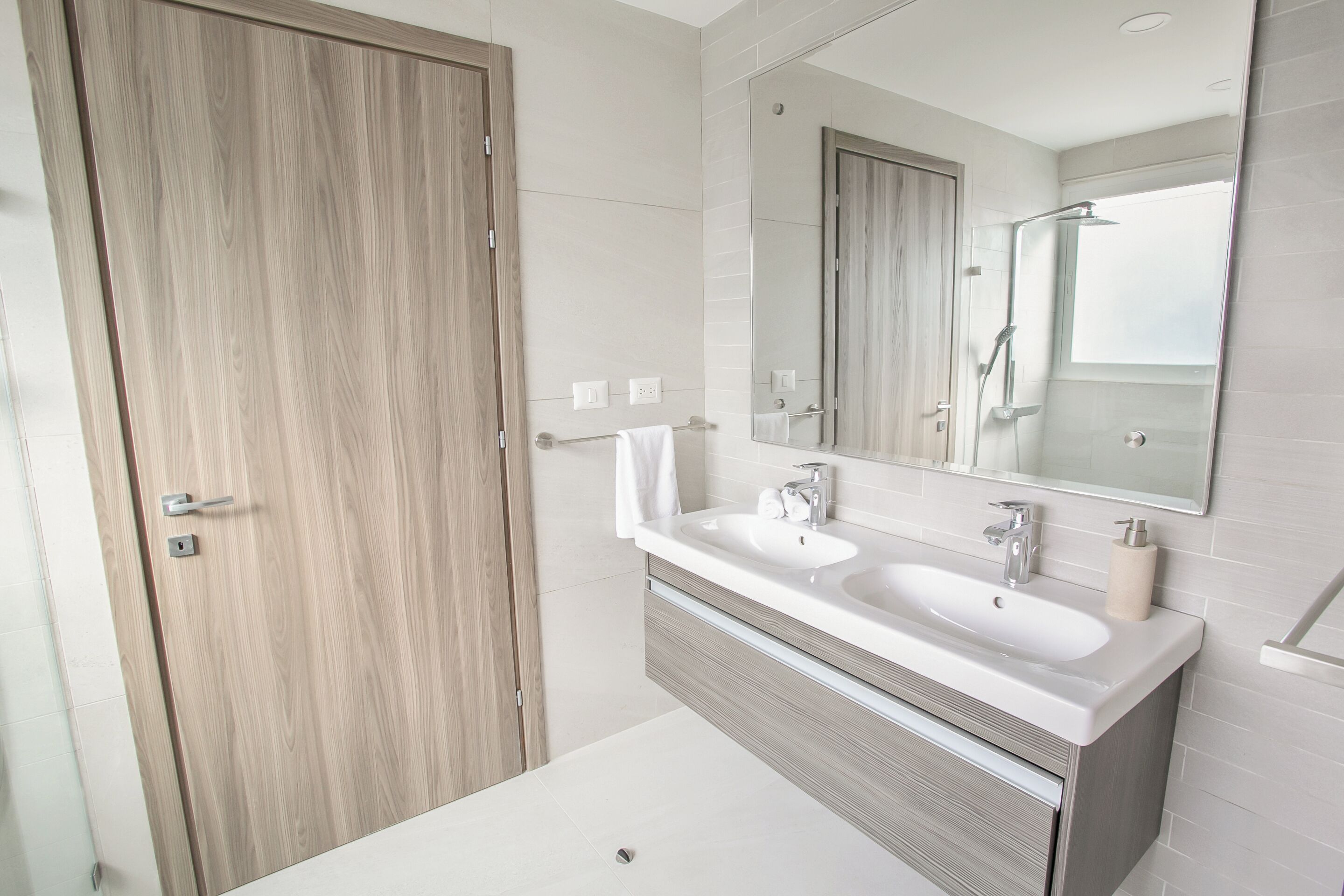 A modern bathroom with a wooden door, a double sink vanity, and a large mirror.