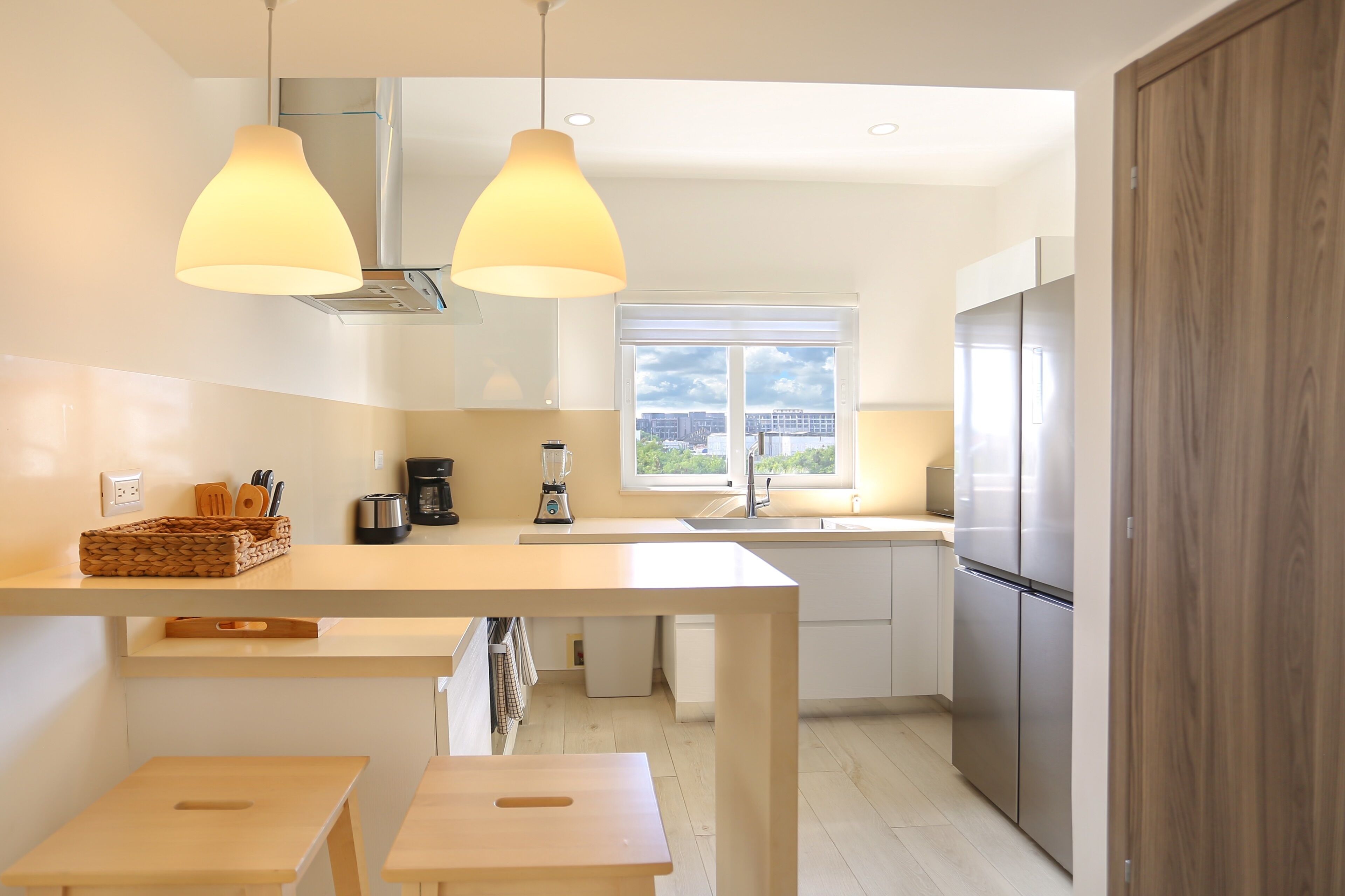 Cozinha moderna com ilha central, luminárias pendentes, janela com vista para a cidade e eletrodomésticos de aço inoxidável.