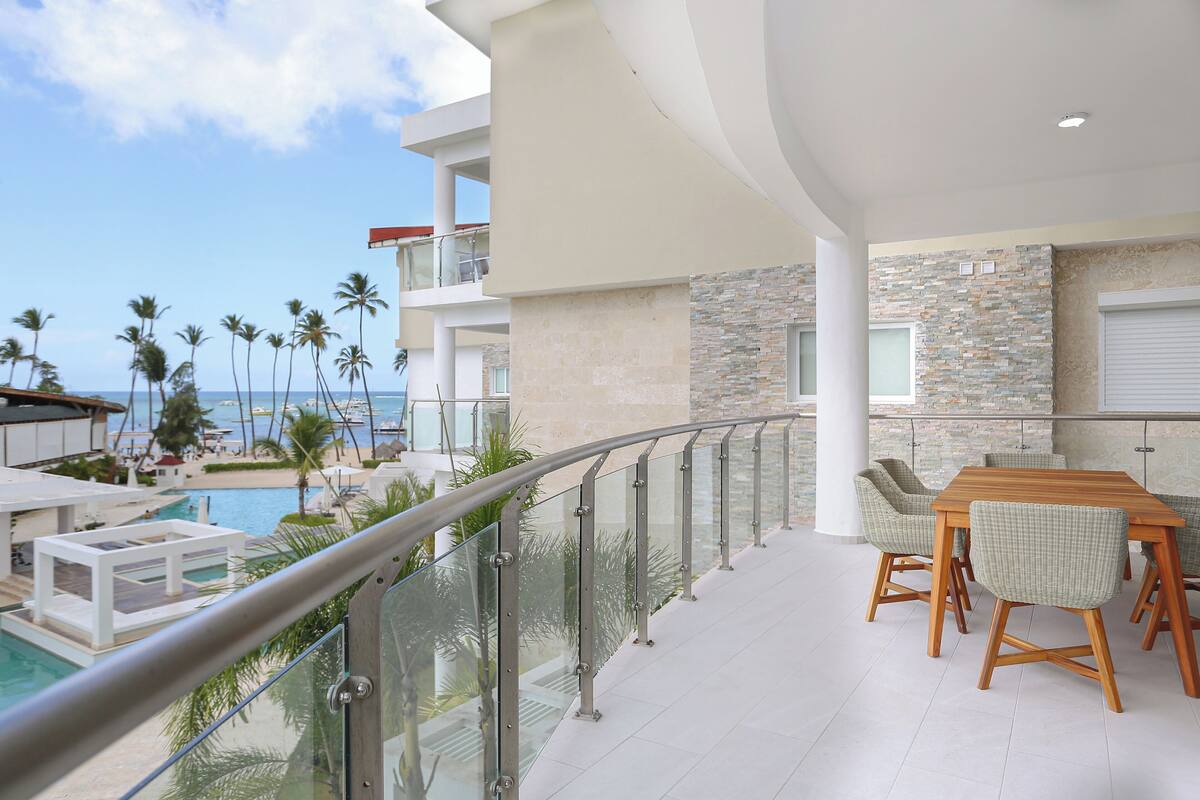 Un balcon avec vue sur une piscine et des palmiers, un coin repas avec des chaises et une table, et un mur en pierre.