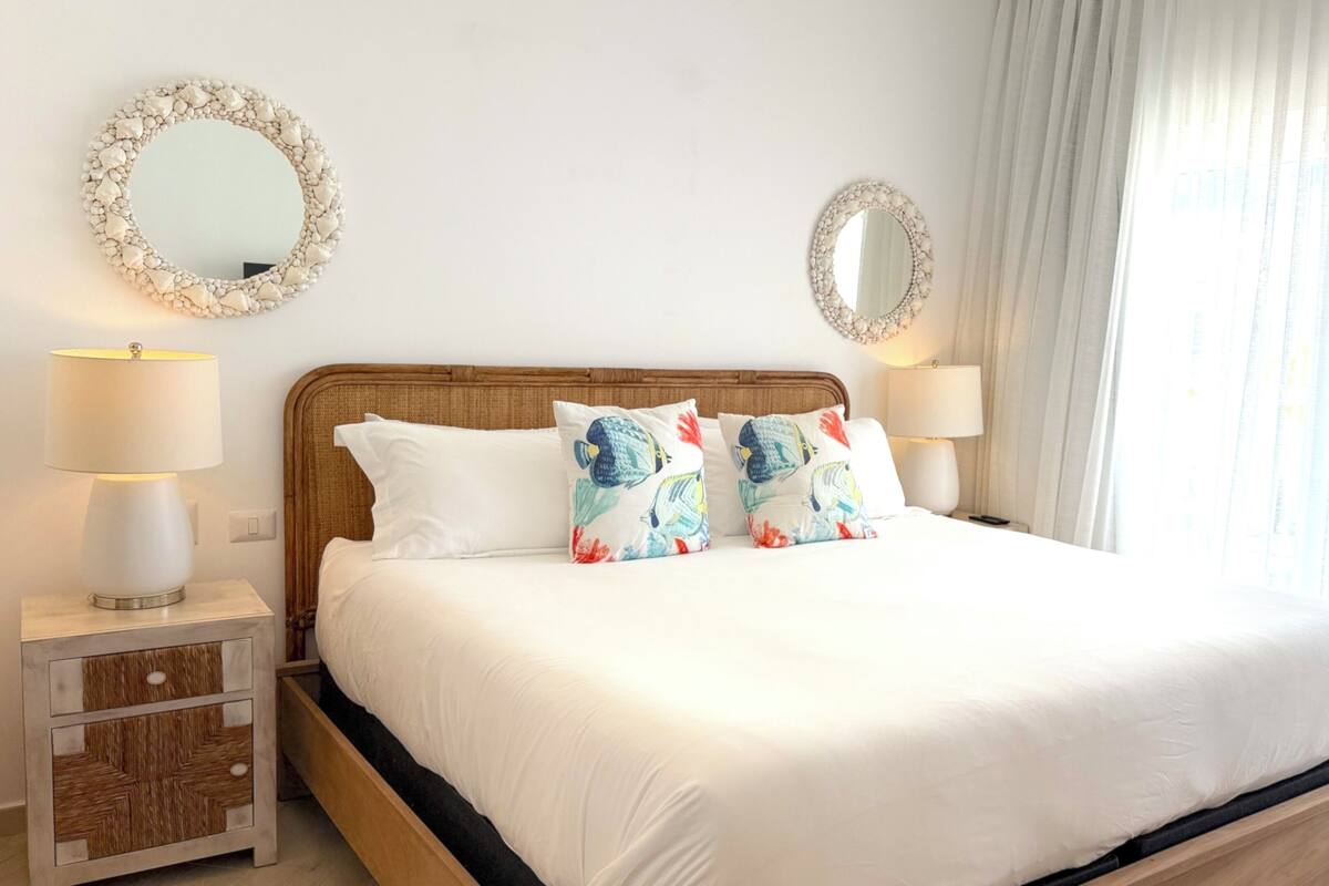 Un lit bien fait, avec une literie blanche, deux coussins décoratifs, une tête de lit en bois et deux tables de chevet avec des lampes.