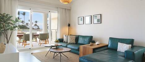 Een moderne woonkamer met groene leren banken, een ronde salontafel en een groot raam met uitzicht op palmbomen.