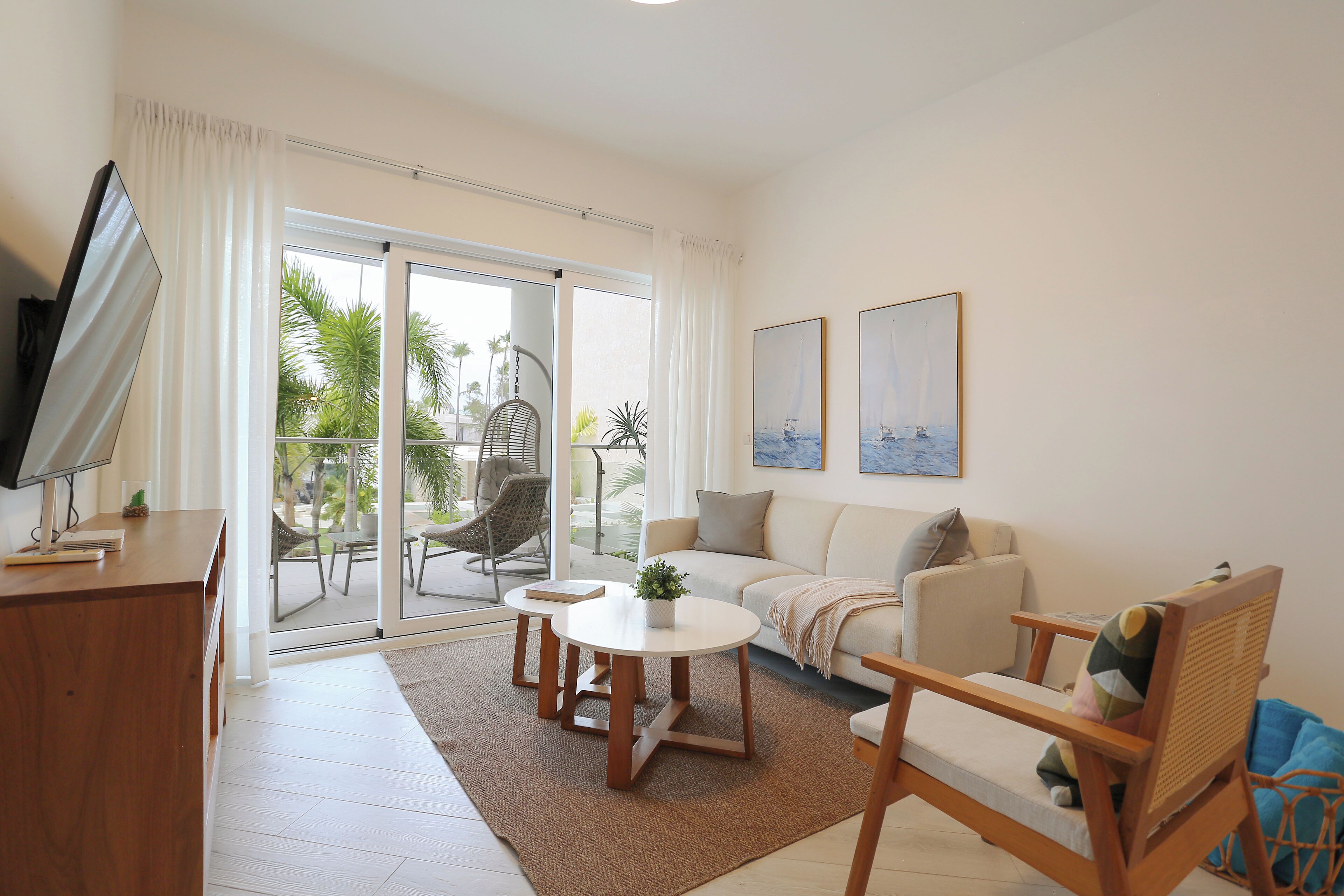 A living room with a sofa, coffee table, and chairs, a television, and a sliding glass door leading to an outdoor area with a chair and plants.