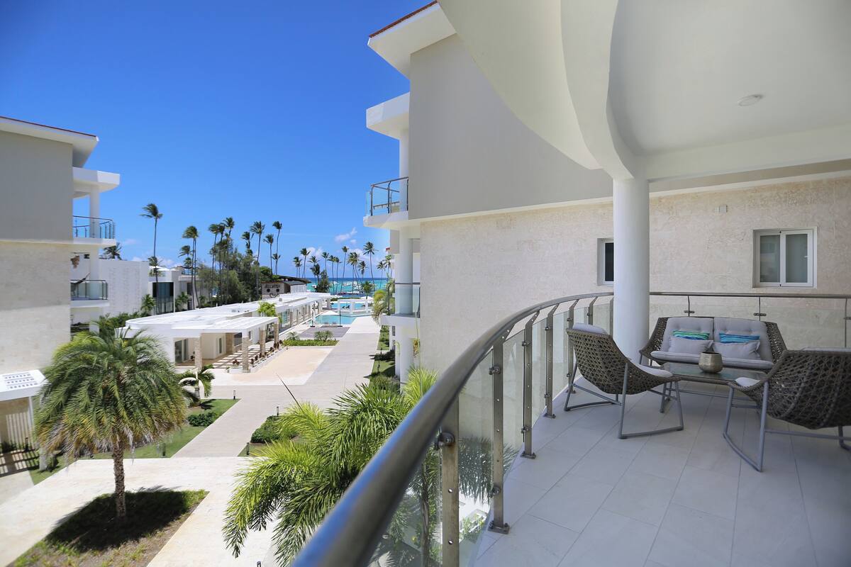 Un balcon d’hôtel avec vue sur la piscine, un espace salon extérieur et des palmiers.