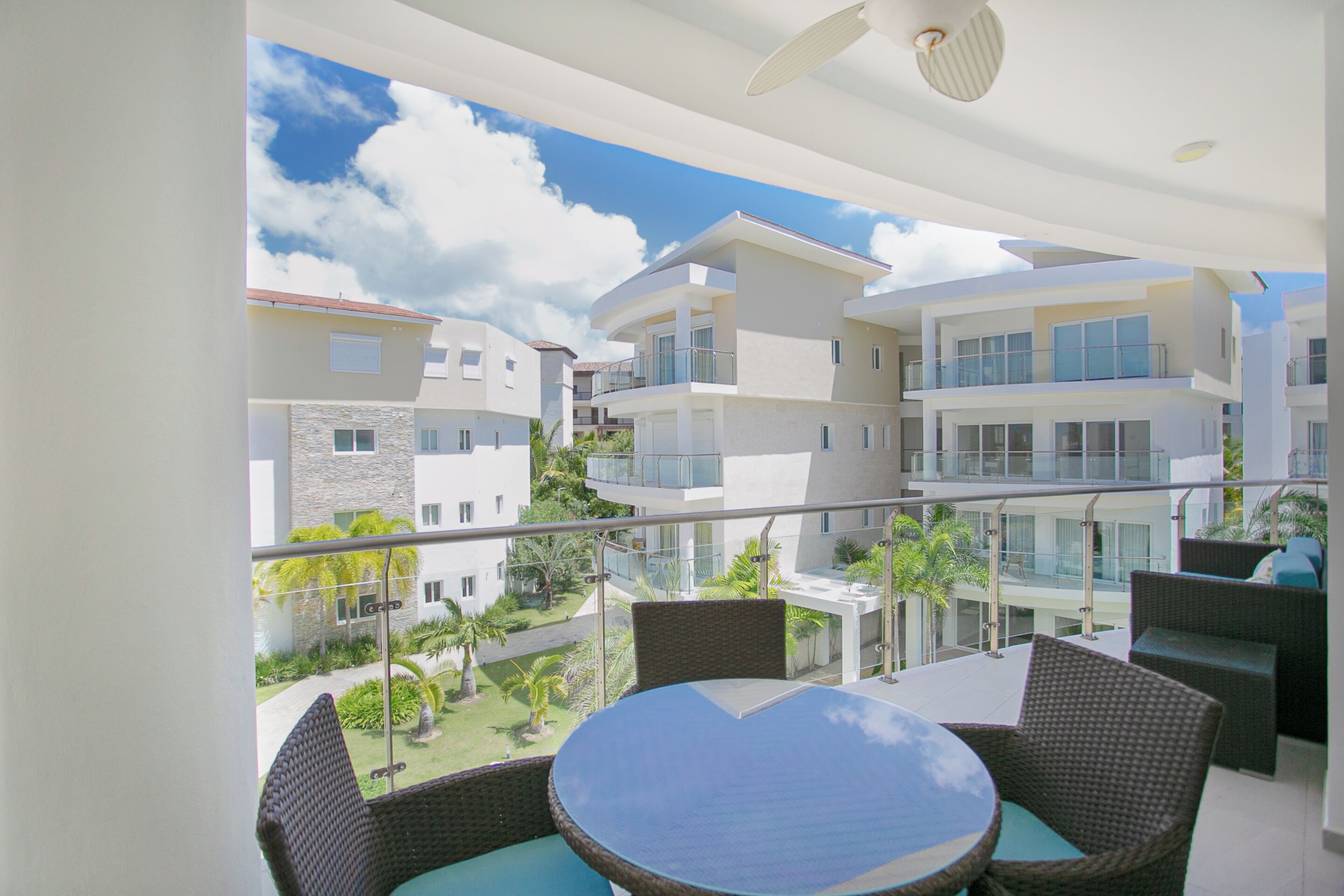 A balcony with wicker furniture and a glass table, overlooking a residential area with modern buildings and a clear sky.
