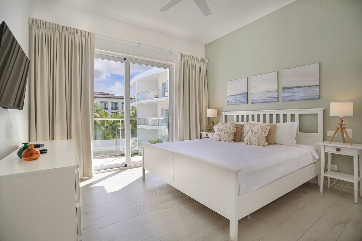 Une chambre à coucher avec un lit, une table de chevet, une lampe, une fenêtre et une vue sur un bâtiment à l’extérieur.