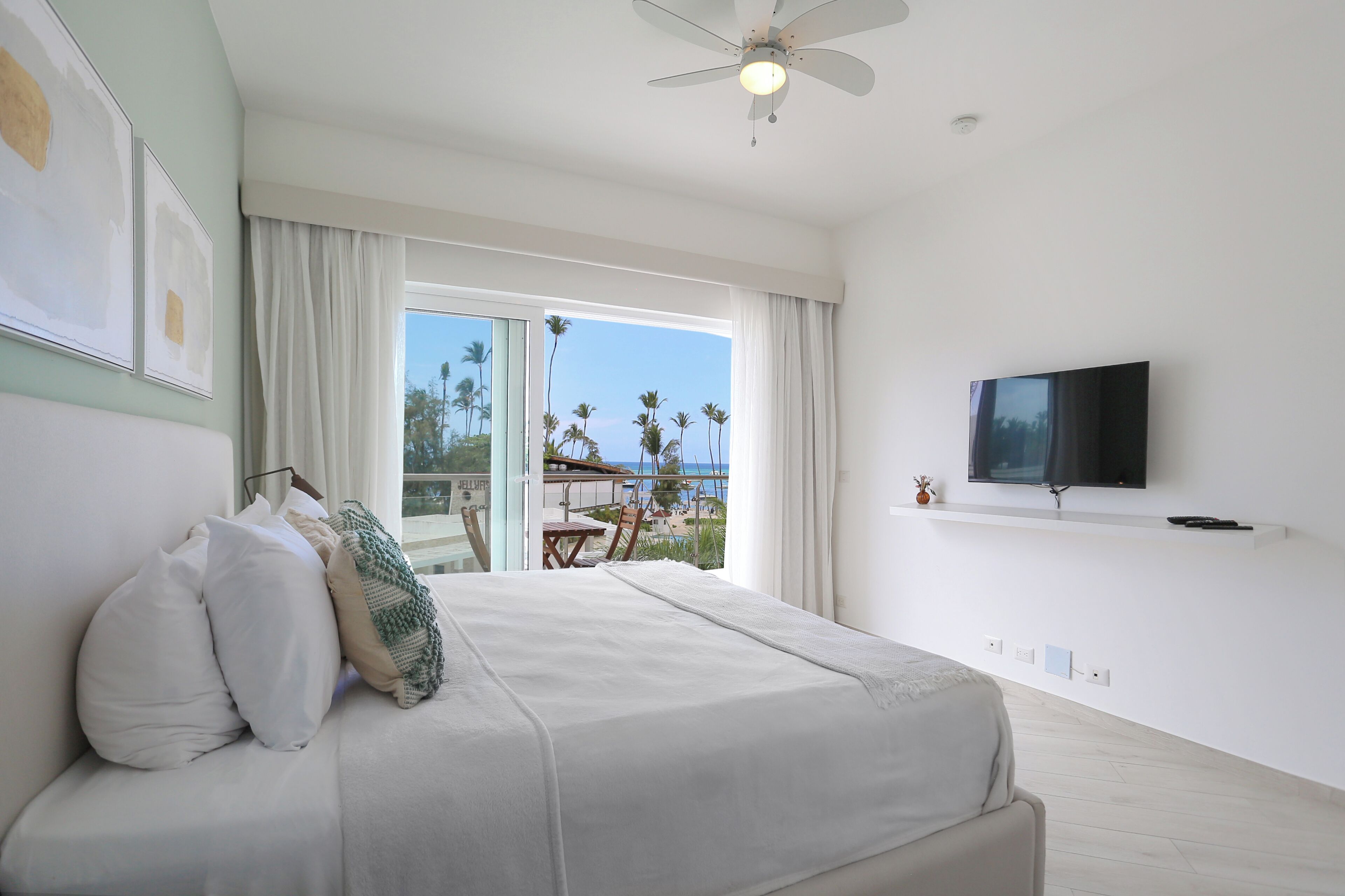 Habitación de hotel con cama, televisor montado en la pared y balcón con vista a palmeras y al océano.