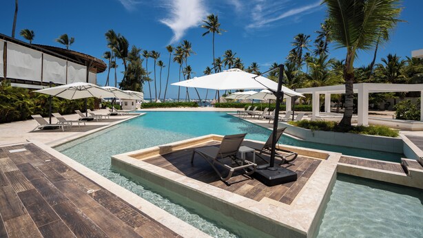 Un espace piscine agrémenté de chaises longues et de parasols, entouré de palmiers et offrant une vue sur un ciel dégagé.
