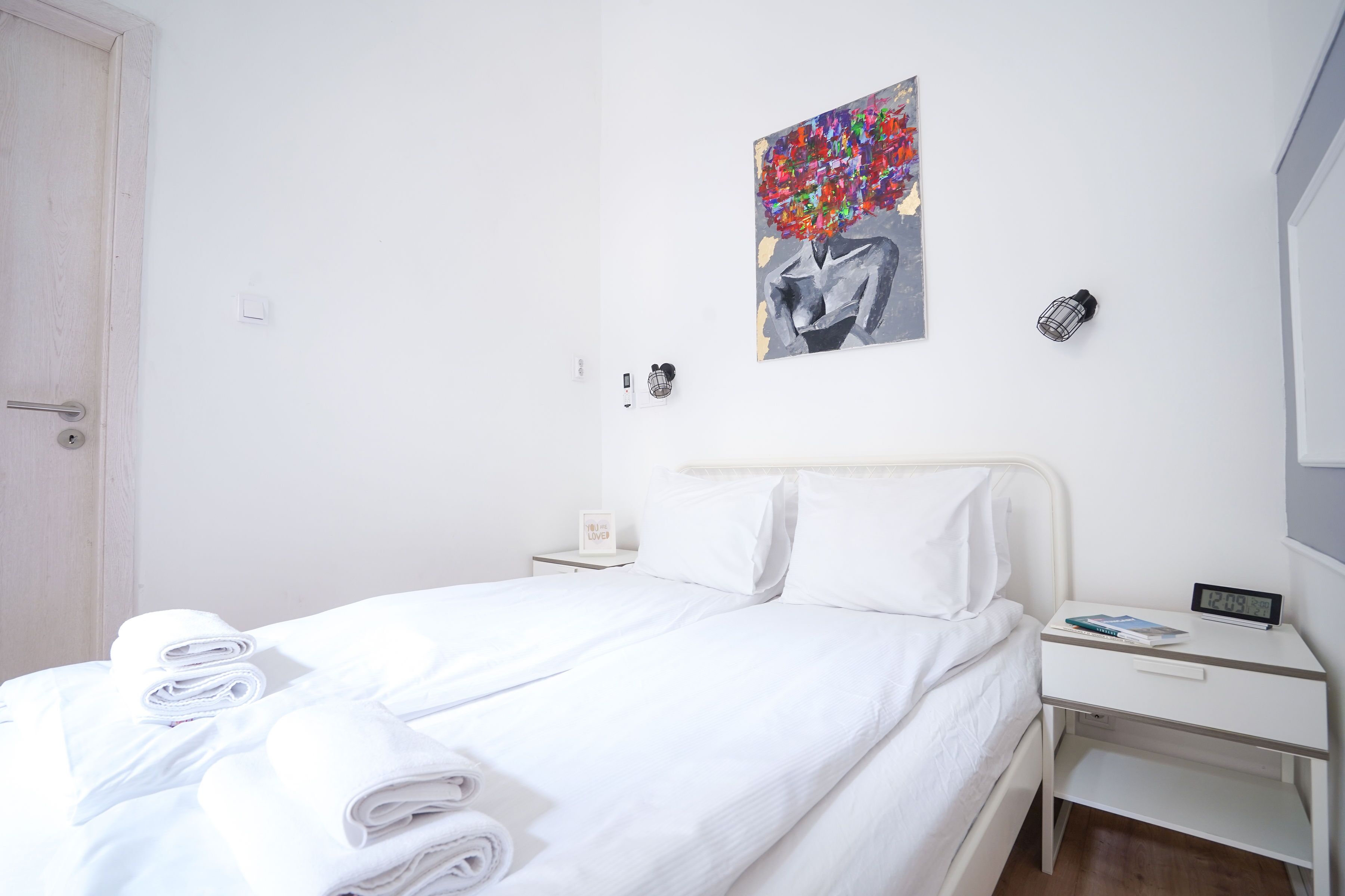 Et hotelværelse med en pænt redt seng, et natbord med en lampe, et vægur og et maleri.