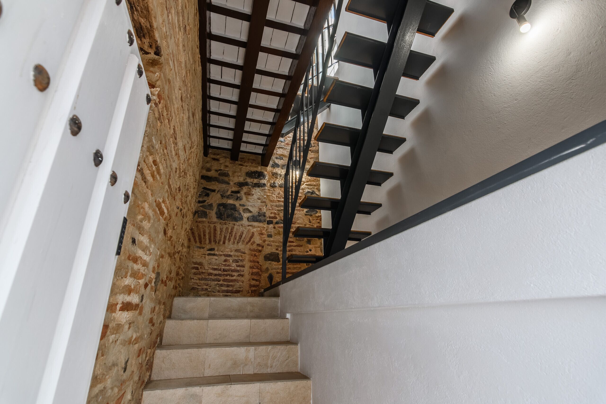 Eine Treppe mit einer Steinmauer an einer Seite und einer weißen Wand an der anderen, die zu einer Holzzieldecke mit einer Leuchte führt.