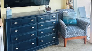Un salon avec un grand téléviseur à écran plat, une commode bleu foncé dotée de nombreux tiroirs, un fauteuil agrémenté d’un coussin sur le thème de la plage, une horloge murale et un panneau indiquant « MAISON DE PLAGE ».