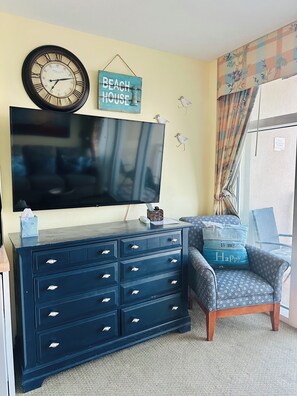 A living room with a large flat-screen TV, a dark blue dresser with multiple drawers, a chair with a beach-themed pillow, a wall clock, and a sign that reads 'BEACH HOUSE'.