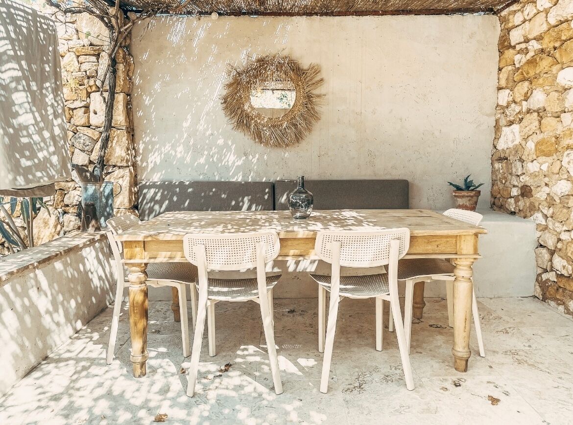 Eine Terrasse im Freien mit einem Holztisch und weißen Korbstühlen, einer Steinwand und einem dekorativen Kranz an der Wand.