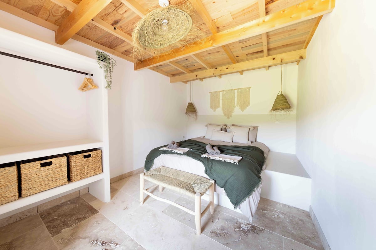 A bedroom with a wooden ceiling, a bed with a green blanket, a bench, and wicker baskets on a shelf.