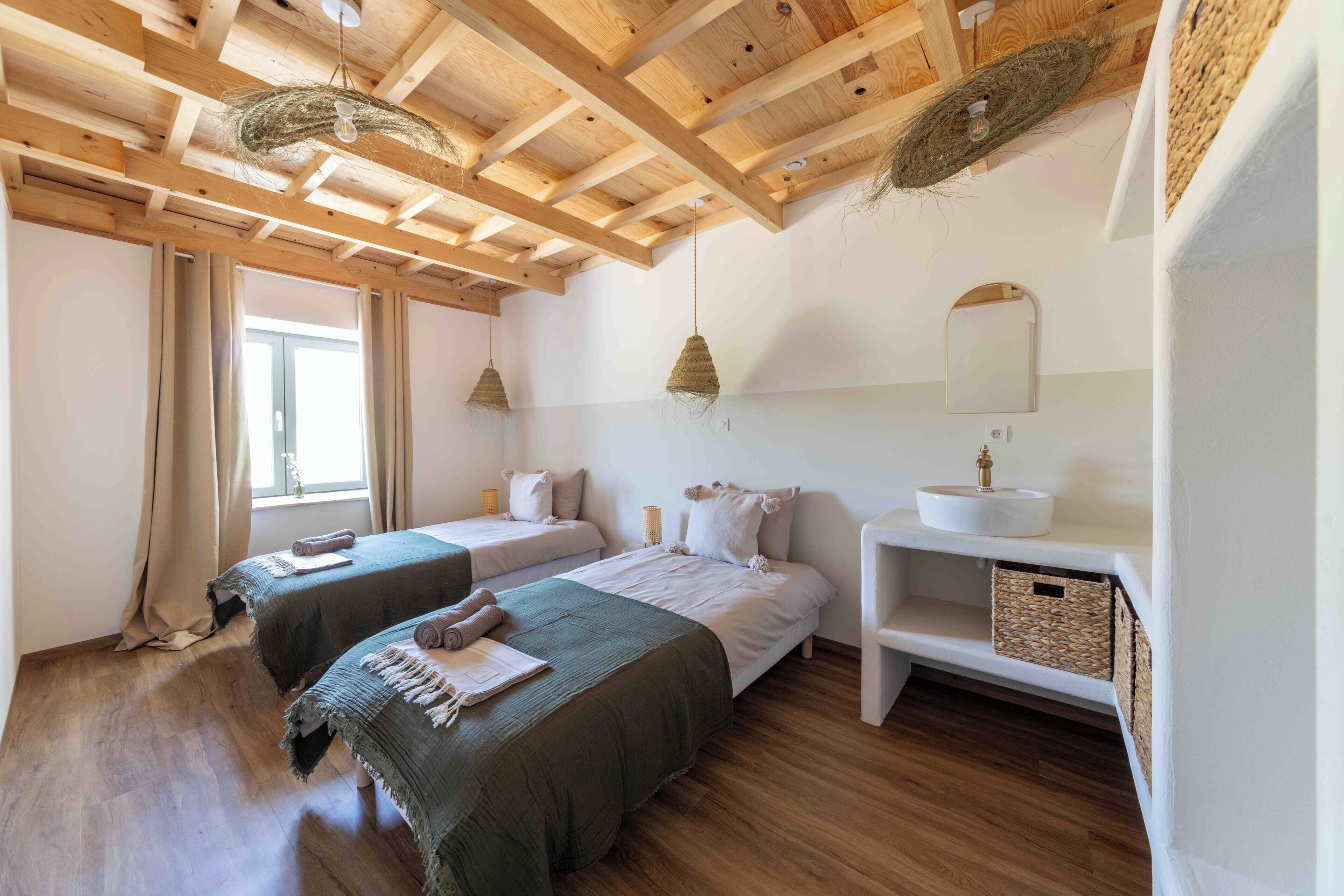 Une chambre avec deux lits, des poutres apparentes en bois au plafond, un lavabo blanc et un miroir.