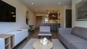 Un soggiorno moderno con un divano grigio, un tavolino da caffè bianco con oggetti decorativi, una TV a parete e un angolo cucina sullo sfondo.