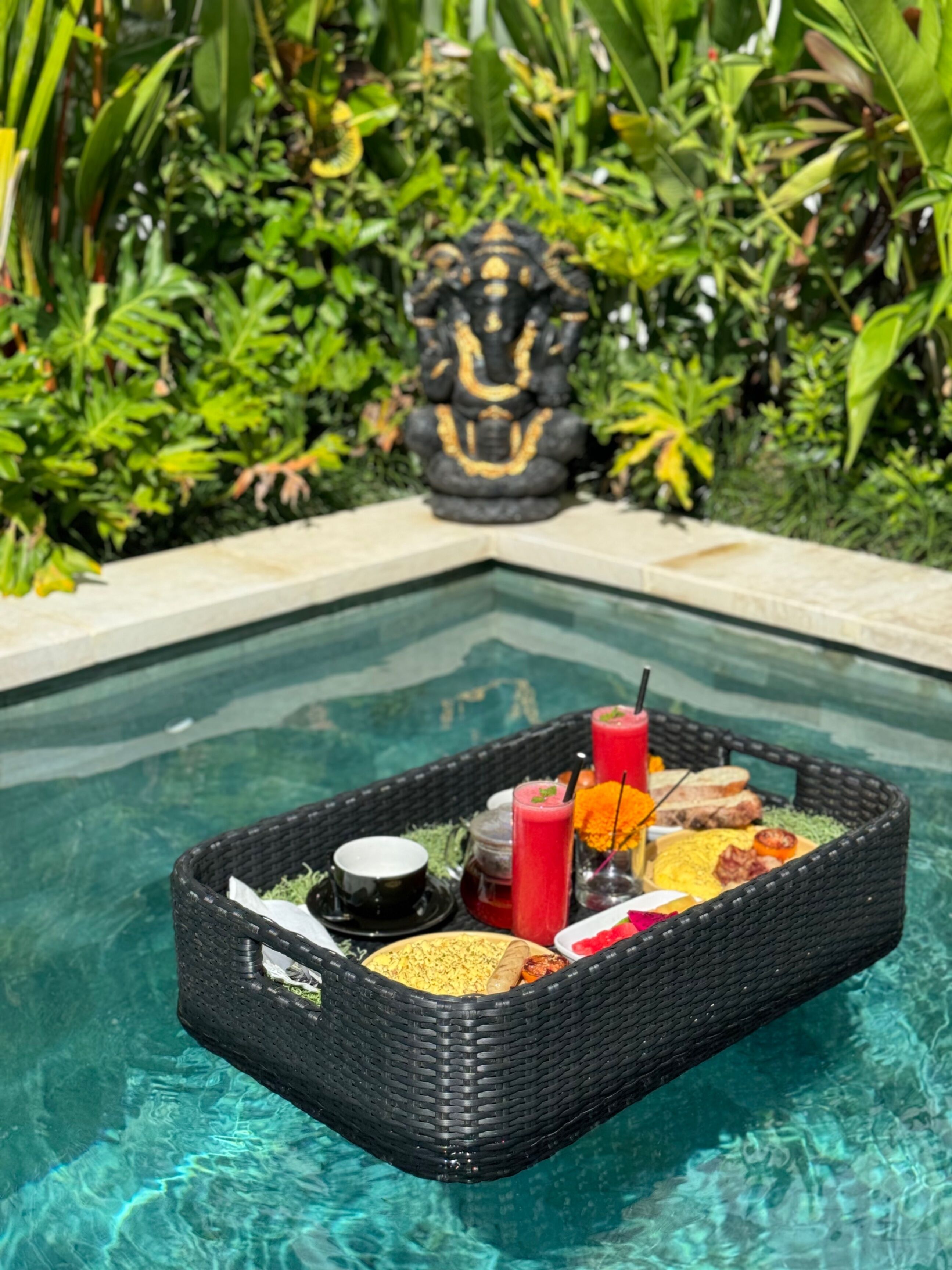 A poolside dining setup with a wicker basket containing food and drinks, surrounded by lush greenery and a decorative statue.