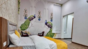 Una cama doble con cabecera de madera, ropa de cama blanca y una manta amarilla. Hay dos cuadros de arte con motivos de pavo real sobre la cama.