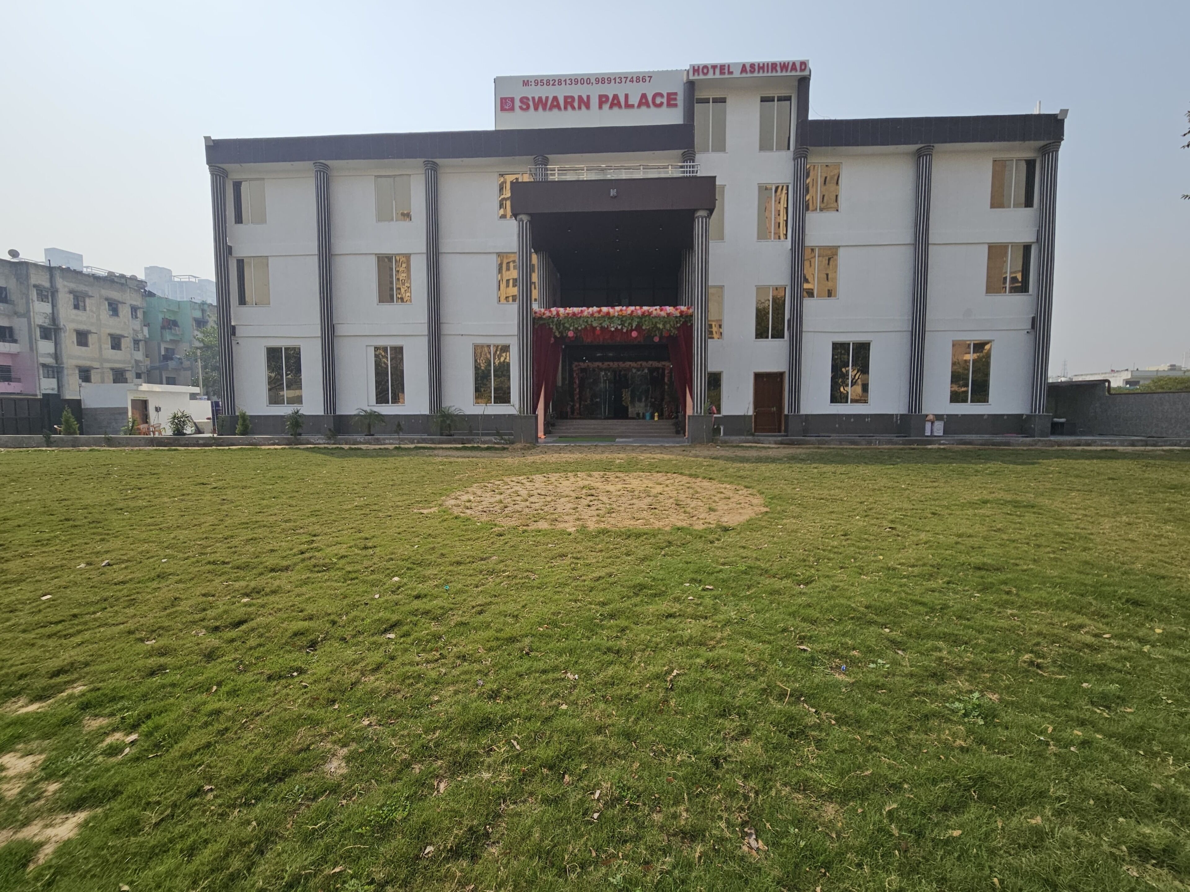 A hotel named Swarn Palace with a central entrance, a lawn in front, and a multi-story building with columns.