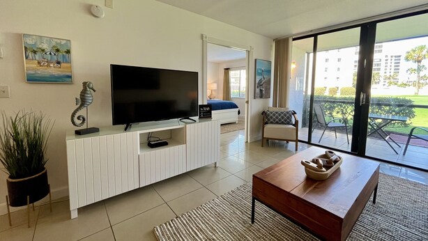 A modern living room with a flat-screen TV, a white entertainment unit, a wooden coffee table, and a large sliding glass door leading to an outdoor patio. - GV 5317 Golf Course View - Welcome To Paradise (Fort Pierce)