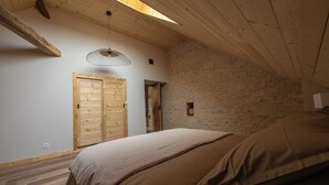 A bedroom with a sloped ceiling, wooden beams, a bed with brown bedding, a wooden wardrobe, and a stone wall. - LE Grenier DU Jean (Sancey)
