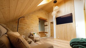 A cozy room with a wooden ceiling, a stone wall, a wooden floor, a sofa, a chair, a TV, and a woven light fixture. - LE Grenier DU Jean (Sancey)