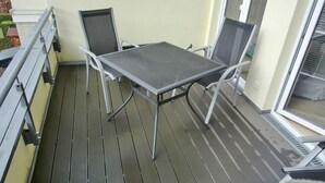 Uma varanda com mesa de metal e quatro cadeiras, corrimão e janela.