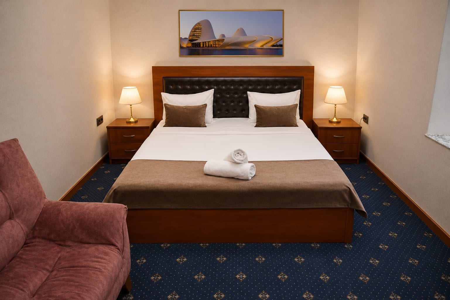 Een hotelkamer met een groot bed, twee nachtkastjes met lampen, een hoofdbord, een schilderij boven het bed en een kamerbreed tapijt met dessin.