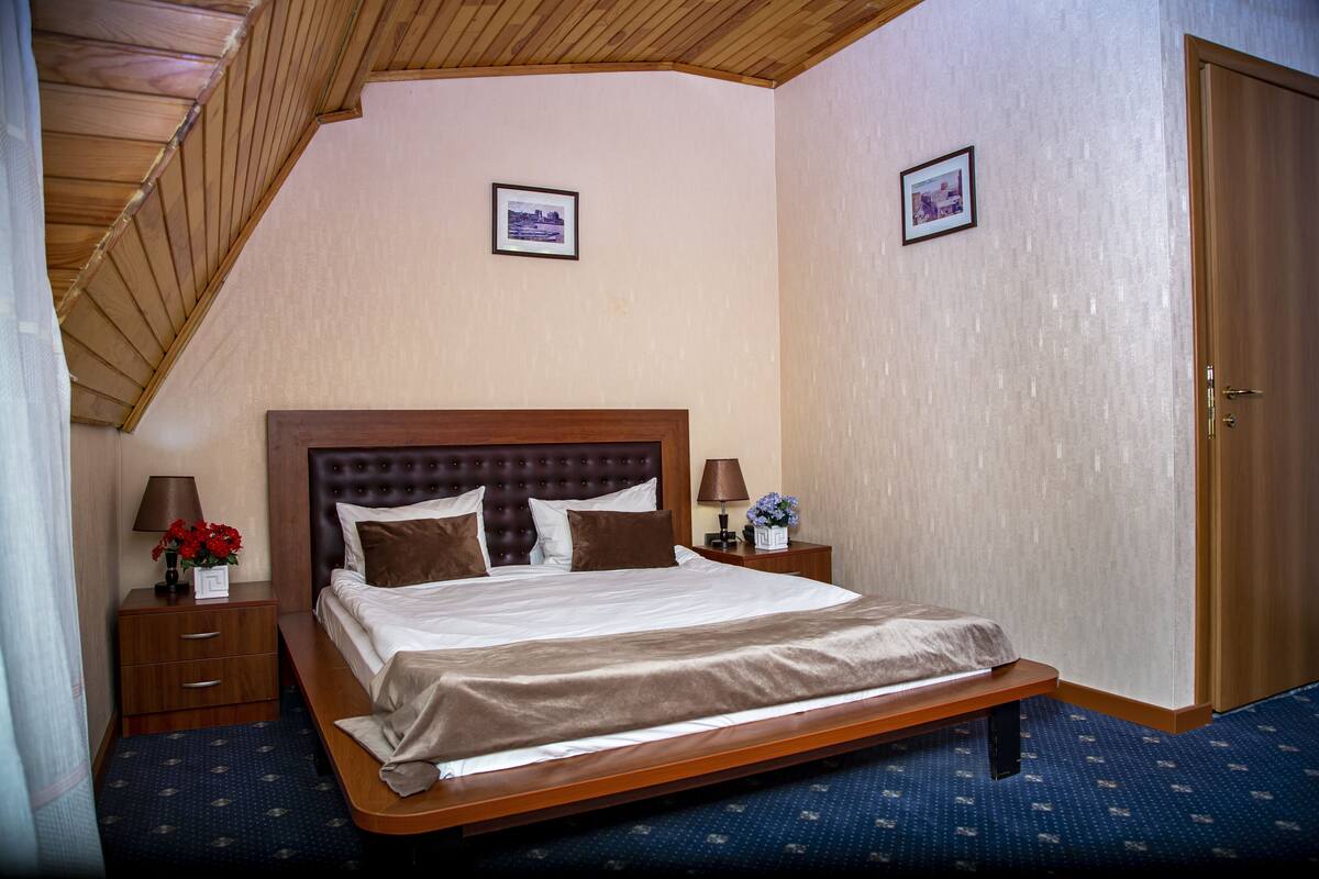 A neatly arranged hotel room with a large bed, wooden headboard, two bedside tables with lamps, and framed pictures on the walls.