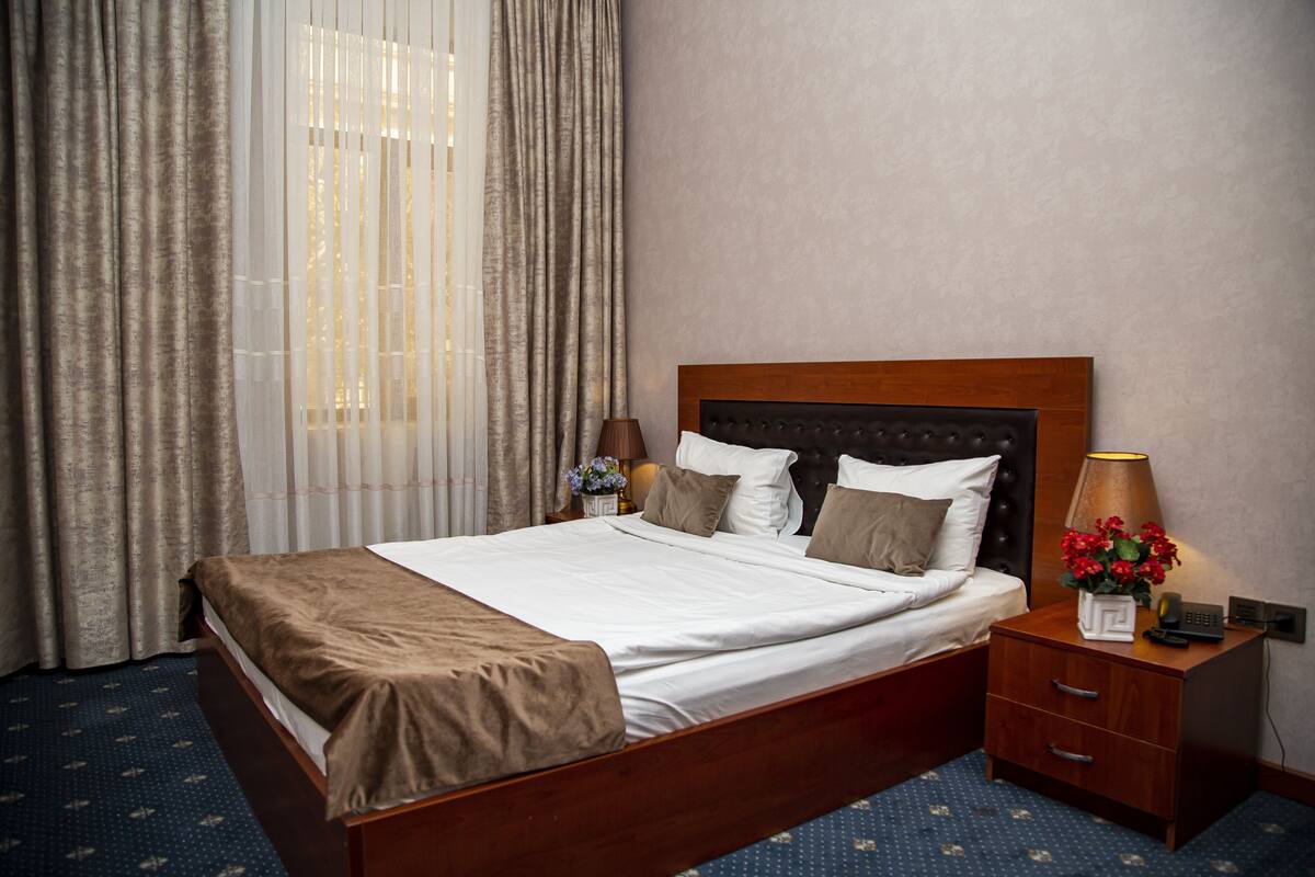 A hotel room with a wooden bed, a nightstand with a lamp, and a view of the outdoors through sheer curtains.