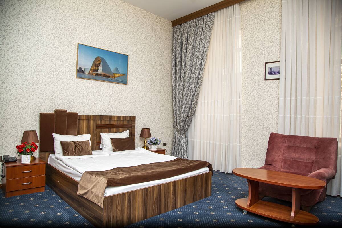 A hotel room with a wooden bed, a red armchair, a small wooden table, a nightstand with a lamp, and a painting on the wall.