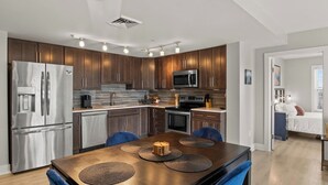 A modern kitchen with stainless steel appliances, wooden cabinets, and a dining area with a wooden table and blue chairs. - Lakeview Condo | Seasonal Hot Tub | Pool | Restaurant (Canandaigua)