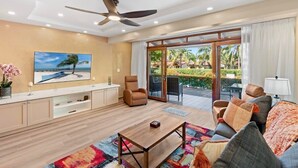 Un salon moderne doté d’un téléviseur à écran plat, d’un ventilateur de plafond, d’un canapé, d’une table basse et offrant une vue sur un jardin tropical grâce à de grandes portes coulissantes.