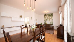 A dining area with a wooden table and chairs, a kitchen in the background, and a living room with a sofa and a plant. - Villa Torchio (Manerba del Garda)
