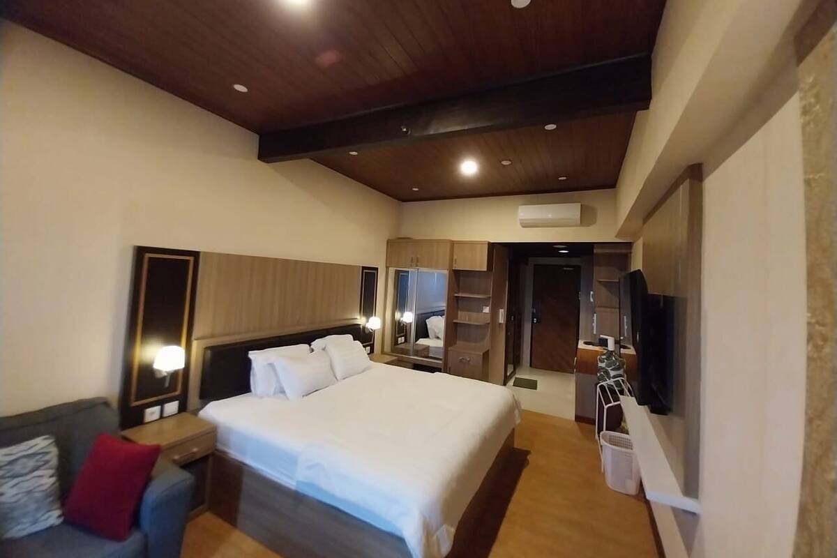 A hotel room with a large bed, a TV mounted on the wall, a wardrobe, and a desk with a lamp.
