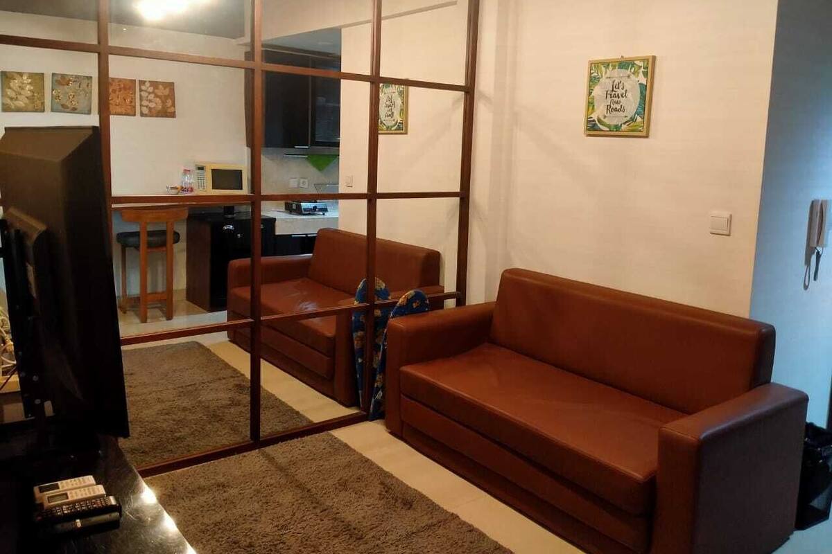 A living room with a brown leather sofa, a television, and a kitchen area visible through a glass partition.