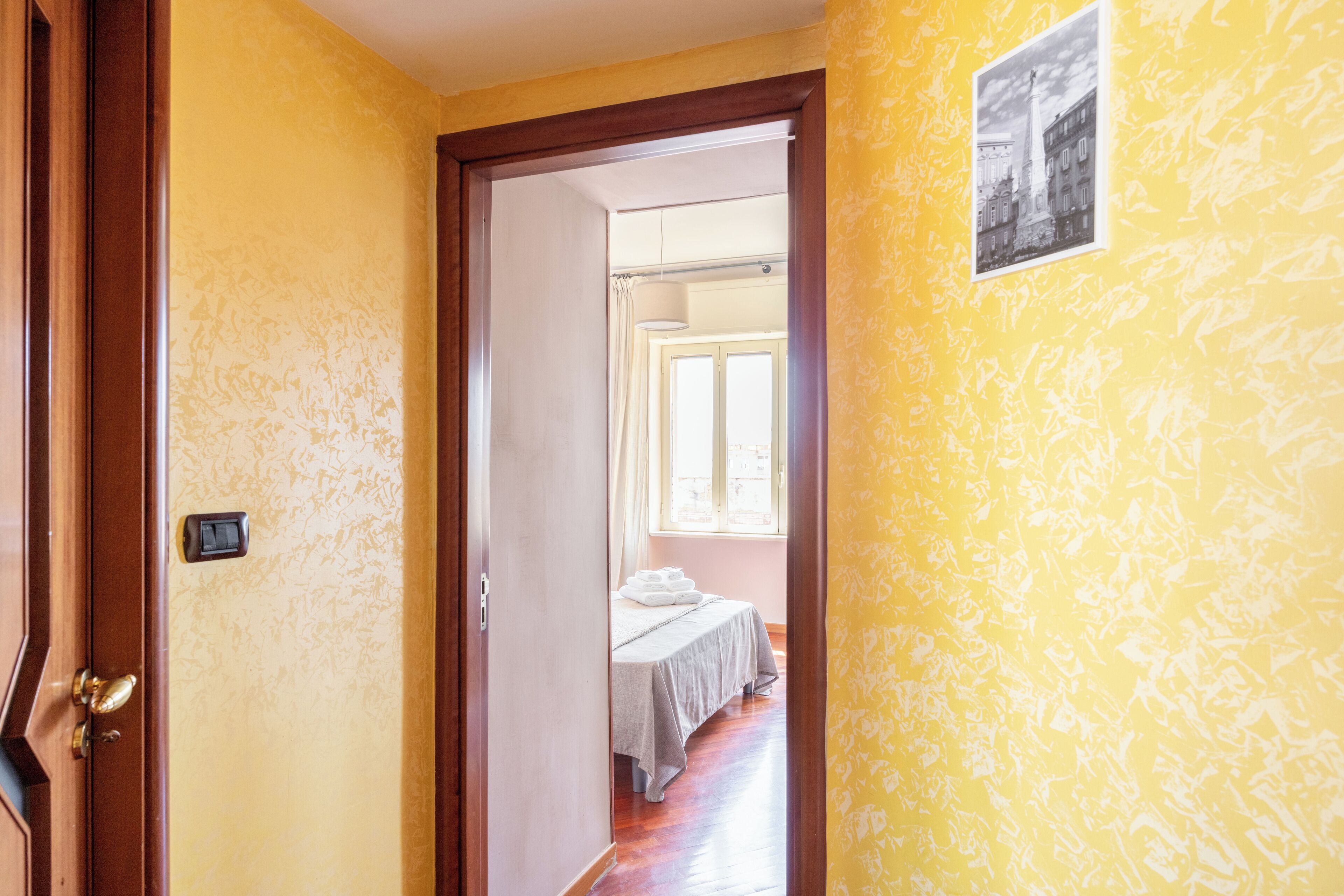 A hallway leading to a room with a bed, a door with a handle, and a wall-mounted light fixture.