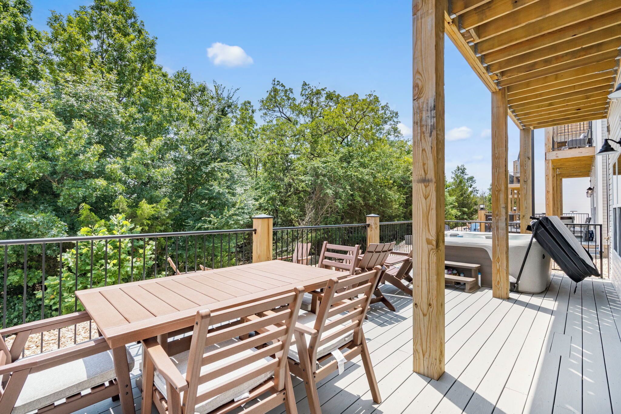 A wooden deck with a table and chairs, surrounded by greenery and a hot tub.