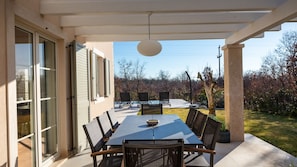 Une terrasse couverte avec une table et des chaises, un luminaire suspendu et une vue sur un jardin et des arbres.