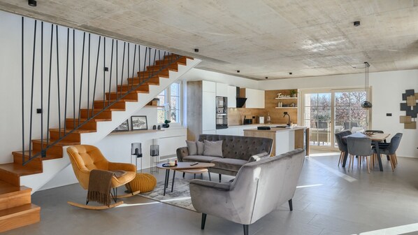 A spacious living area with a staircase, modern furniture, and a kitchen in the background.