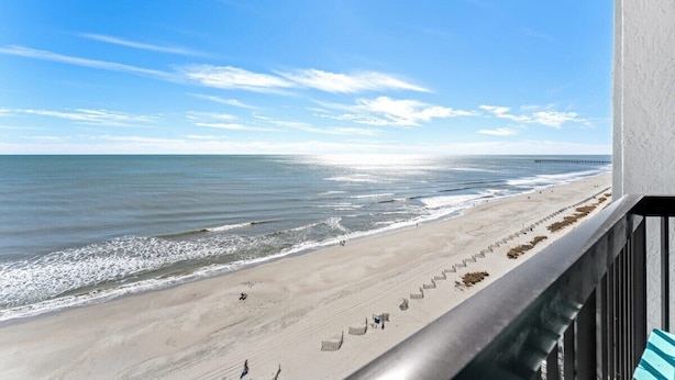 Strandudsigt fra en balkon med gelænder, hav og en mole i det fjerne.