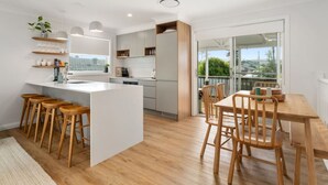 A modern kitchen with a central island, wooden stools, and a dining area with a table and chairs. - Four bedroom  in Australia, Anna Bay (Anna Bay)