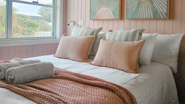 A bedroom with a large window, a bed with multiple pillows, a blanket, and two wall-mounted lamps. - Four bedroom  in Australia, Anna Bay (Anna Bay)