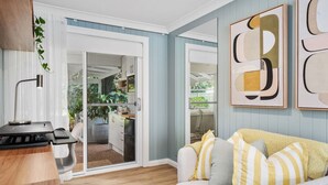 A modern living room with a wooden desk, a grey sofa with striped cushions, and a sliding glass door leading to a kitchen. - Four bedroom  in Australia, Anna Bay (Anna Bay)
